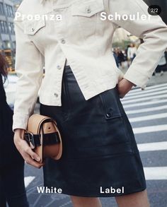 a woman in a white jacket and black skirt is holding a brown purse while standing on the street