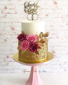 a wedding cake decorated with pink roses and gold leaves on a white table next to a brick wall