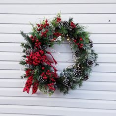 a christmas wreath hanging on the side of a white building with pine cones and red berries