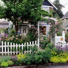 a white picket fence in front of a house with lots of flowers and trees around it