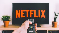 a hand holding a remote control in front of a television with the word netflix on it