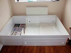 an empty bed frame with magazines on the bottom and side drawers underneath it, in front of a window