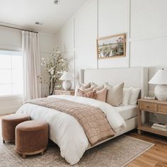 a bedroom with white walls and wood flooring has a large bed, two stools, and a painting on the wall