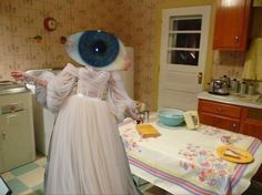 a woman in a white dress with an eyeball on her head standing next to a table