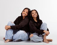 two women are sitting on the floor posing for a photo together, both wearing jeans and black shirts