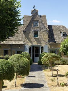 a stone house with trees and bushes around it's front door, on a sunny day