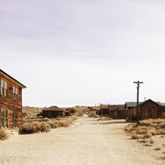 an empty dirt road in the middle of some small buildings and telephone poles on either side