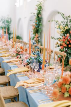 the table is set with blue linens, orange and pink flowers, gold place settings, and candles