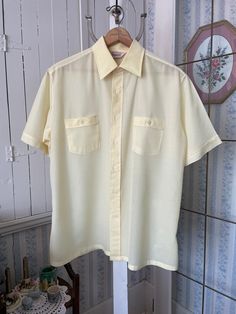 This is a very light weight, semi sheer dress shirt from Kingsport, made from 100 per cent polyester in pale buttery yellow (it's a bit darker than in the photos). The shirt has the original buttons on the front and two chest pockets with matching buttons. Marked size large. The measurements, taken with the shirt lying flat, are: shoulder to shoulder, 19 inches; armpit to armpit, 23 inches; sleeves, 10 1/2 inches; length, 31 inches; bottom edge, 26 inches. In very good condition. Light Yellow Button Up Shirt, Collared Cream Shirt For Summer, Classic Summer Shirt For Daywear, Cream Collared Shirt For Summer, Summer Cream Collared Shirt, Cream Short Sleeve Shirt With Button Closure, Cream Short Sleeve Workwear Shirt, Solid Vintage Summer Shirt, Beige Short Sleeve Shirt For Daywear