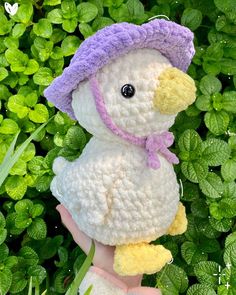 a crocheted stuffed duck wearing a purple hat and scarf in front of green leaves