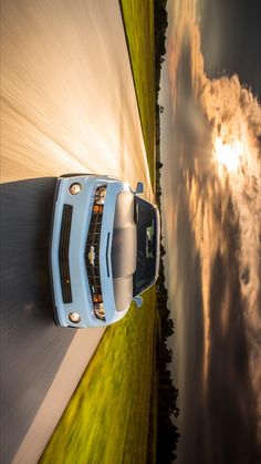 a blue sports car driving down a road next to grass and clouds in the sky