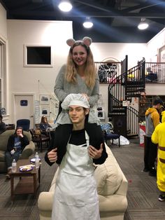 a woman standing next to a man in an apron on top of his head and another person behind him