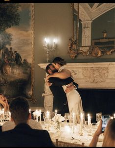 a bride and groom hug in front of candles at their wedding reception with guests seated around the table