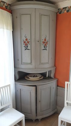 a corner cabinet with painted flowers on the doors and two white chairs in front of it