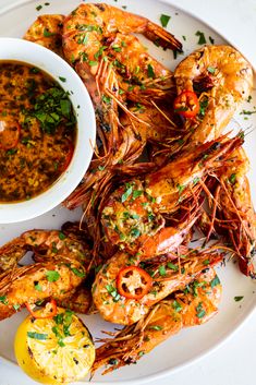 a white plate topped with cooked shrimp next to a bowl of sauce and lemon wedges