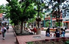 people are sitting on benches in the middle of a city street with shops and trees