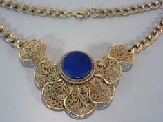 A wonderful vintage necklace that has a beautiful stationary filigree pendant with a round cobalt blue glass disc in the center.  The chain has a decorative pattern and the metal looks to be brass and possibly Czechoslovakian origin. A collar length necklace measuring 16 inches long, the center adornment is 3 1/2 inches of that length and drops down about 2 inches. In very good condition, may want to clean/polish the metal. To see all the jewelry in my Etsy shop, go to my home page here ~ www.et Antique Gold Filigree Round Necklace, Antique Gold Filigree Necklaces, Antique Gold Round Filigree Necklace, Ornate Blue Necklace With Intricate Design, Antique Blue Jewelry With Filigree, Antique Blue Filigree Jewelry, Antique Blue Brass Necklace, Vintage Blue Filigree Jewelry, Blue Metal Filigree Jewelry