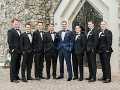 a group of men standing next to each other in front of a stone building wearing tuxedos