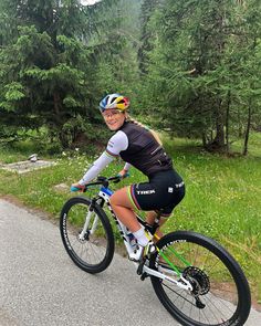 a woman riding a bike down a road next to some green grass and pine trees