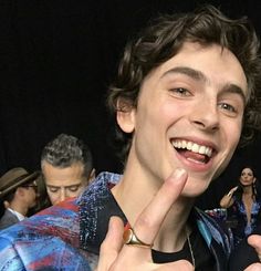 a young man is smiling and giving the peace sign with his hand while standing in front of a group of people