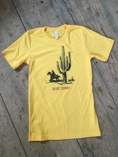 𝔻𝕖𝕤𝕖𝕣𝕥 ℂ𝕠𝕨𝕓𝕠𝕪 Tee Yellow Short Sleeve T-shirt With Screen Print, Mustard Short Sleeve T-shirt With Graphic Print, Unisex Yellow Crew Neck T-shirt, Fitted Yellow T-shirt With Graphic Print, Gold Graphic Tee With Short Sleeves, Gold Short Sleeve Graphic Tee, Gold Short Sleeve Cotton Shirt, Gold Cotton Short Sleeve Shirt, Yellow Relaxed Fit Pre-shrunk T-shirt