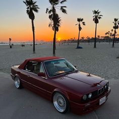 a red car parked on top of a sandy beach next to palm trees at sunset