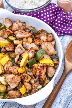 a white bowl filled with chicken and veggies next to two wooden spoons