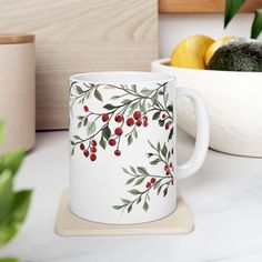 a white coffee mug with red berries and green leaves on it next to a bowl of fruit