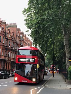 a red double decker bus driving down a street next to tall brown buildings and trees