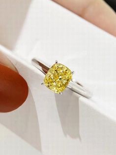 a woman's hand holding a yellow diamond in a white square ring on top of a box