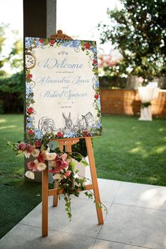 a welcome sign with flowers on it in front of some grass and trees at a wedding