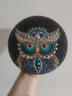 a hand holding a black and blue owl with pearls on it's face, in front of a white wall