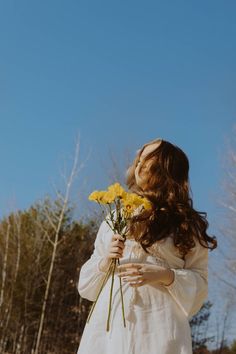 a woman in white dress holding yellow flowers and looking up at the sky with trees behind her