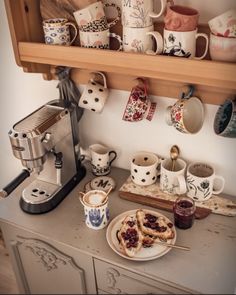 there are many cups and mugs on the shelf above the coffee maker, which is also filled with pastries