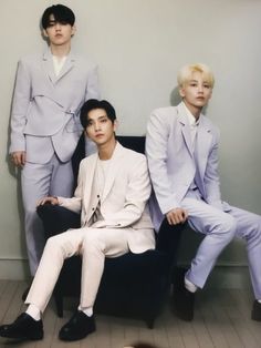 three men in suits are sitting on a black chair and one man is wearing a white suit