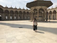 a woman standing in front of a building with arches and pillars on both sides, looking at the ground