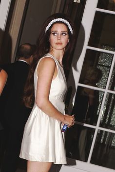 a woman in a short white dress standing next to a window with her hand on her hip