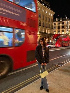 a woman is standing on the sidewalk in front of a red double - decker bus