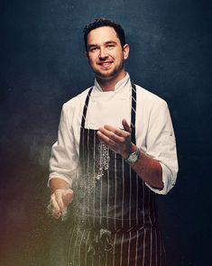 a man wearing an apron and holding his hands out to the side while standing in front of a dark background