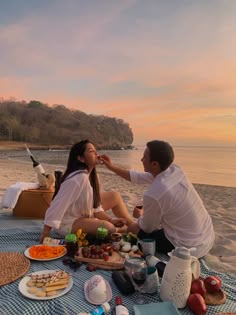 a man and woman sitting on the beach with food in front of them at sunset