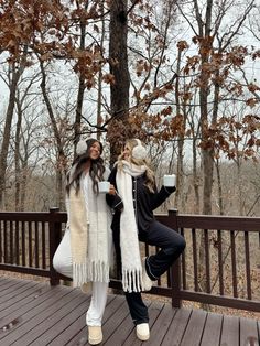 two women standing on a wooden deck in front of a tree and holding coffee cups