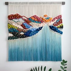 a wall hanging made out of different colored yarns and beads with a plant next to it