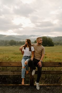 a man and woman sitting on a bench in the middle of a field with mountains behind them