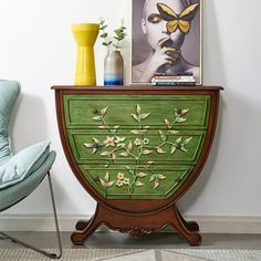 a green cabinet with flowers painted on it next to a blue chair and yellow vase