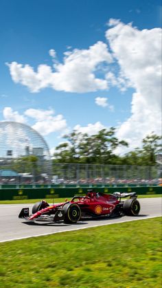 a red race car driving down a track in front of a large green field with white clouds