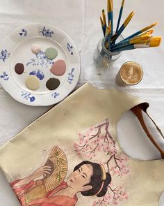 a painting on a table next to some paintbrushes and a plate with an image of a woman holding a fan