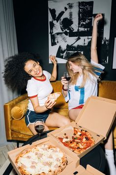 two women sitting on a couch eating pizza and drinking wine