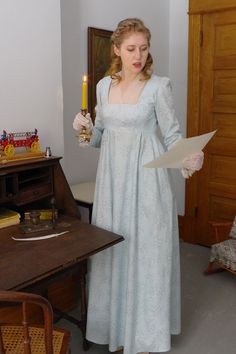 Model is holding a lighted candlestick and is reading a paper standing next to a drop down desk. Corduroy Dress, Historical Dresses