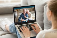 a woman sitting on a couch using a laptop computer with an elderly couple on the screen