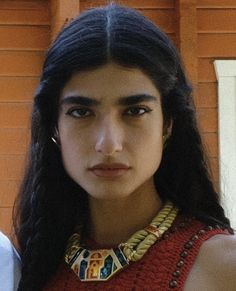 a woman with long black hair wearing a red dress and gold necklace, standing in front of a brick wall